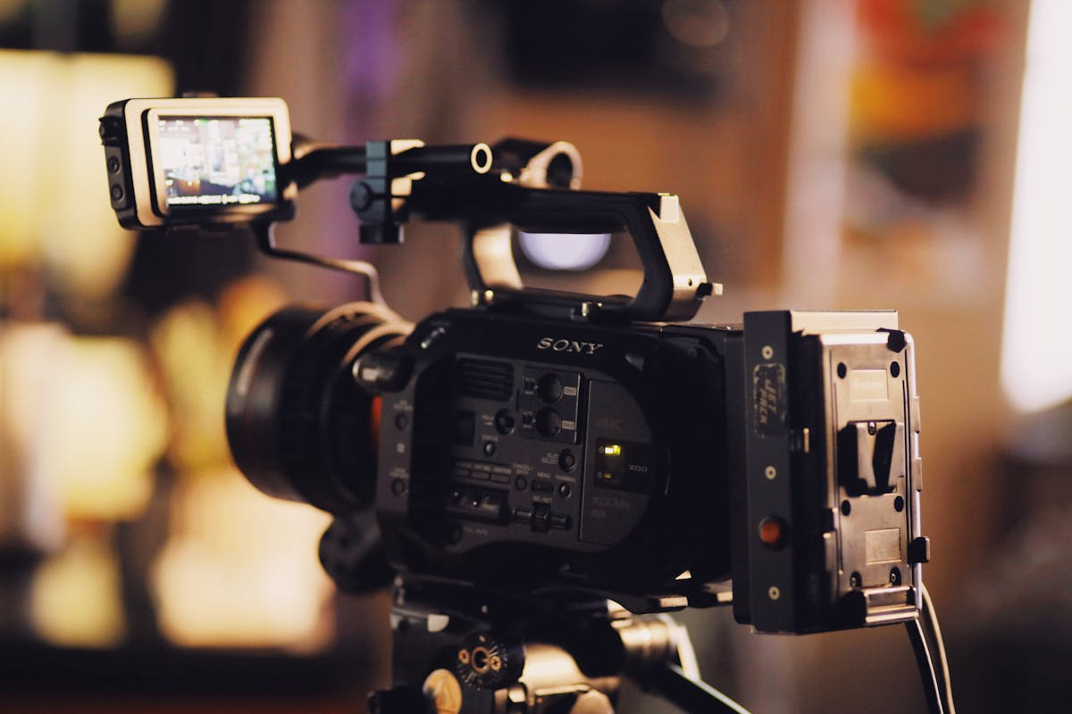 Professional cinema camera with a top-mounted monitor on a tripod in a warmly lit, blurred studio