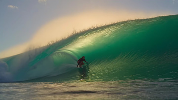 Seedance 2.5 sample: A surfer threads a glowing turquoise barrel at sunset as spray explodes behind her.