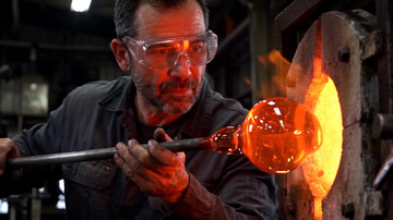Imagera Warlord Reference sample: A glassblower in safety glasses turns a molten gather at the mouth of the furnace.