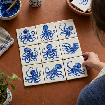 A hand places a painted tile into a grid of tiles showing the same blue octopus.