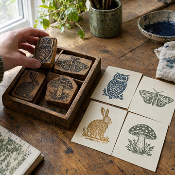 A hand lifts a carved wooden stamp from a tray of custom character stamps beside their inked prints.