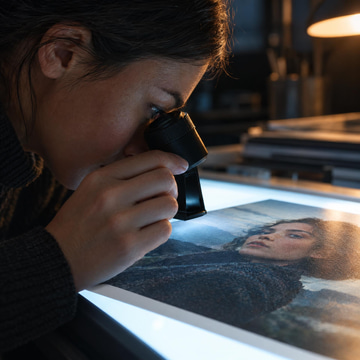 A photo editor studies a printed portrait through a jeweler's loupe on a glowing lightbox.