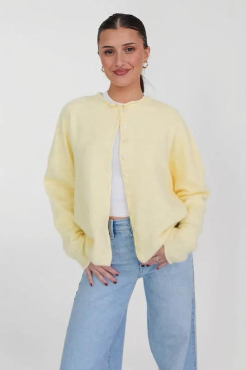 Body photo: a woman with slicked-back hair in a pale yellow cardigan and wide-leg jeans, standing against a white studio wall