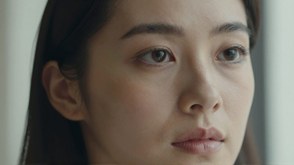 Tight close-up of a young woman's face with dark hair, brown eyes and natural skin texture in soft light