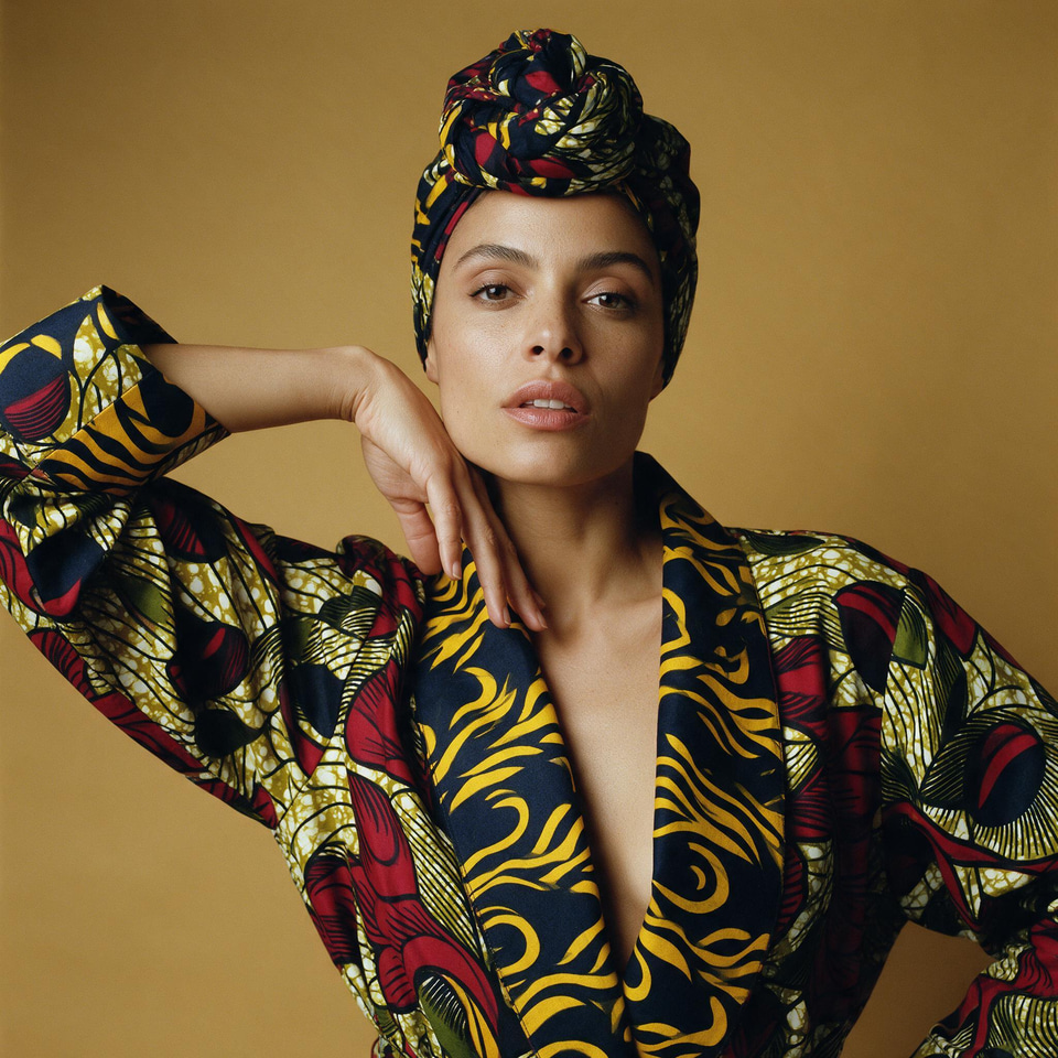 A woman in a bold patterned wrap and headscarf posing against a mustard wall