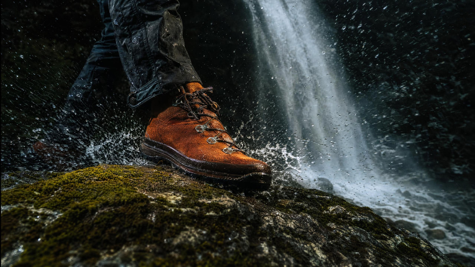 A hiker's rust suede boot slams onto a mossy rock in the rain