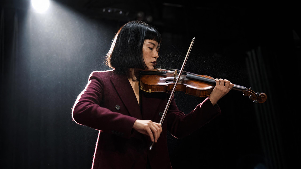 A violinist with a blunt black bob in a burgundy coat plays mid-phrase on an empty stage u