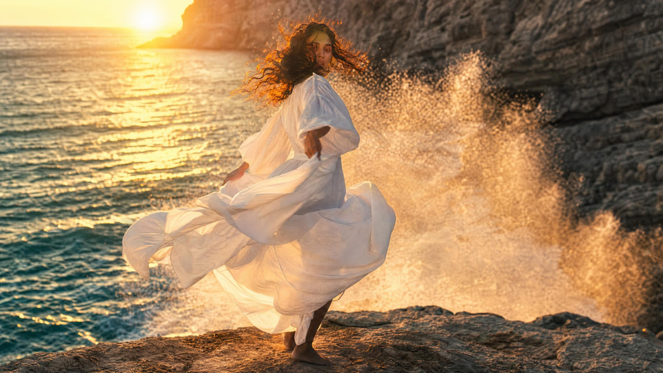 A woman in a white linen dress spins on a cliff edge above the sea at golden hour