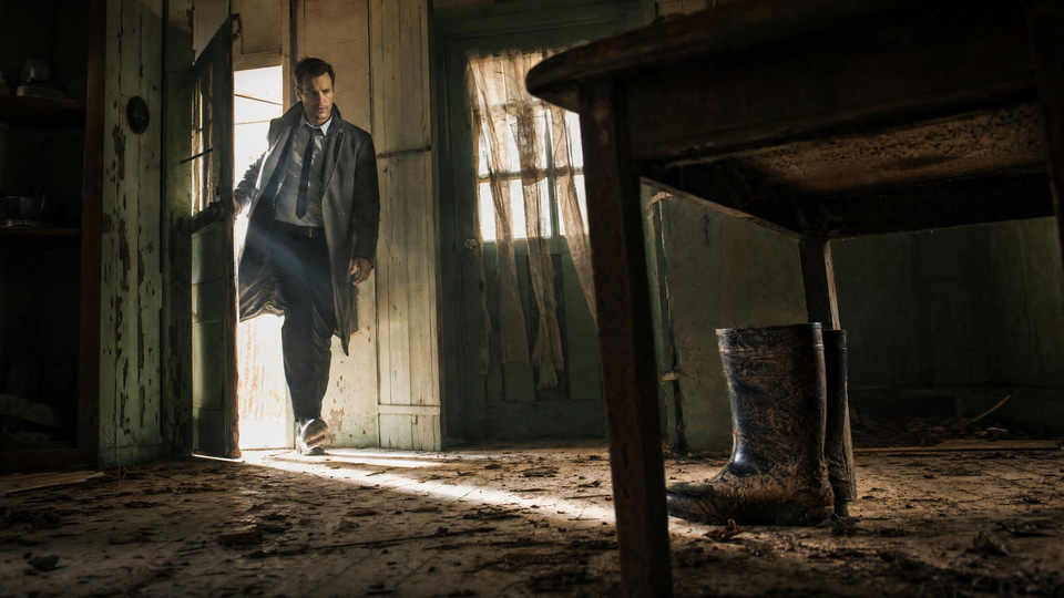 A detective in a long grey coat bursts through the door of an abandoned farmhouse